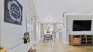 A modern living room with a wooden floor, a TV on the wall, and a dining area in the background. - Calming 3 bedroom home with designer details in Na (Nampa)