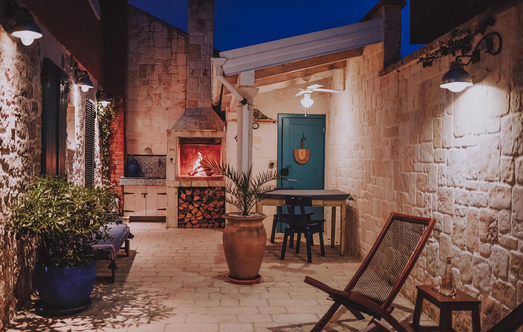 A rustic outdoor patio with a brick wall, a stone fireplace, a wooden table, and a blue door.