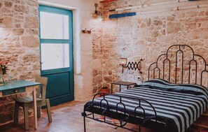 A bedroom with a stone wall, a bed with a metal frame, a small table with a chair, and a window with a teal door. - Gorgeous home in Preko with WiFi (Preko)