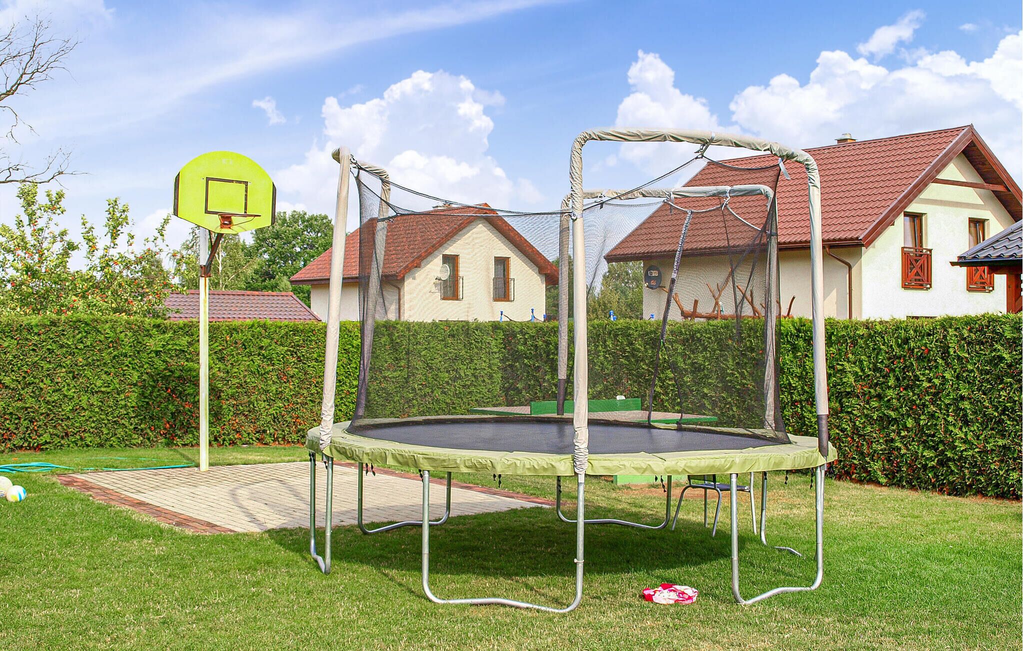 A trampoline in a backyard with a basketball hoop, surrounded by a hedge and houses in the background.