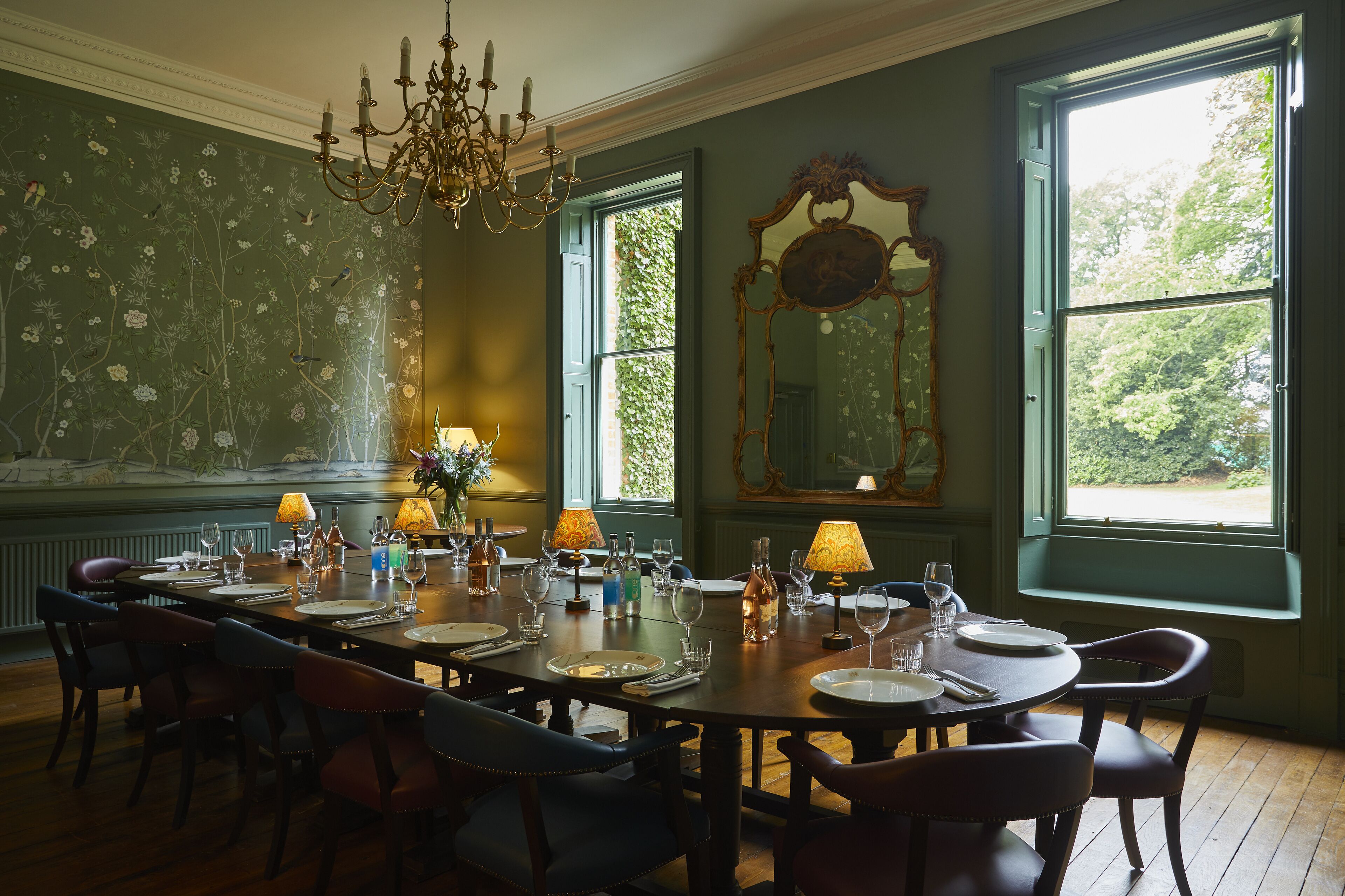 A dining room with a large table set for six, surrounded by chairs, with a chandelier and floral wallpaper.