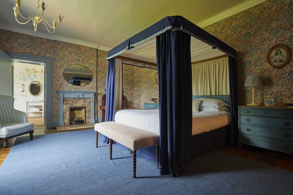 A four-poster bed with a bench, a fireplace, a blue armchair, and a dresser with a mirror.