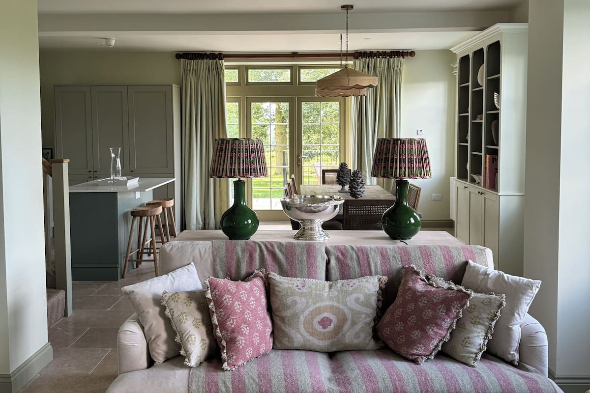 A living room with a sofa, a patterned throw blanket, and a dining table with chairs. There is a kitchen area in the background with cabinets and a window.