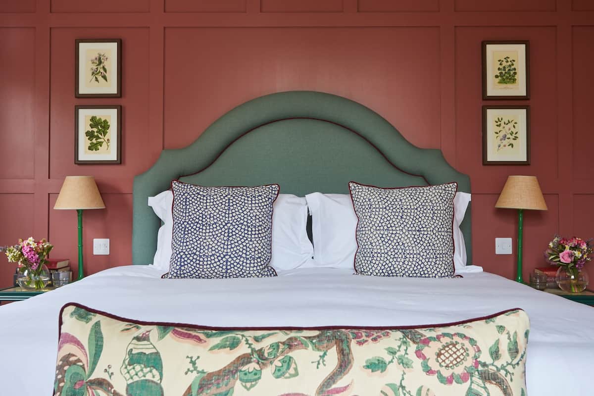 A neatly made bed with patterned pillows, a green upholstered headboard, and framed botanical prints on the wall.