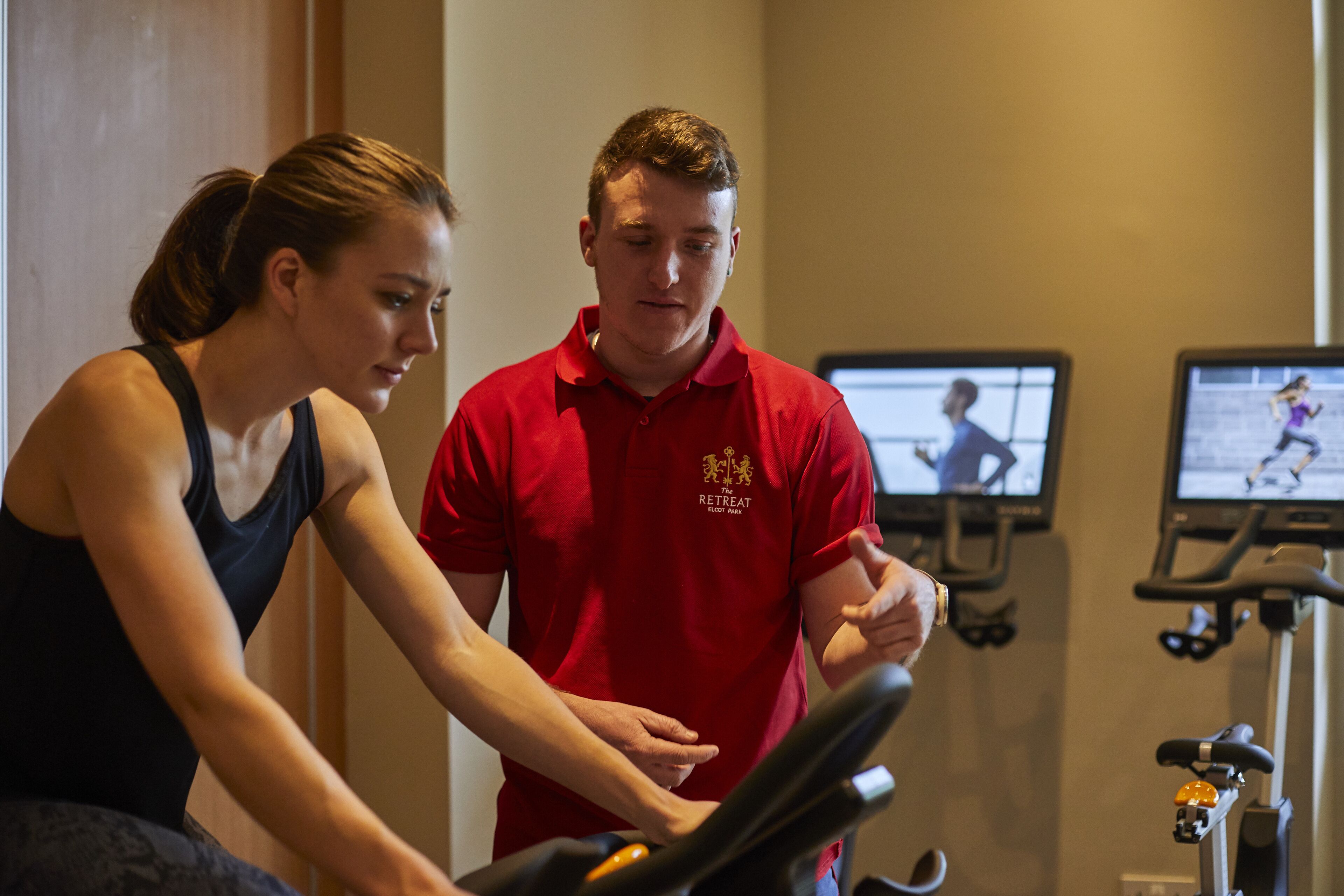 Un hombre con una camisa roja ayudando a una mujer en una caminadora dentro de un gimnasio con equipo de ejercicio y una pantalla de televisión mostrando un programa de entrenamiento.