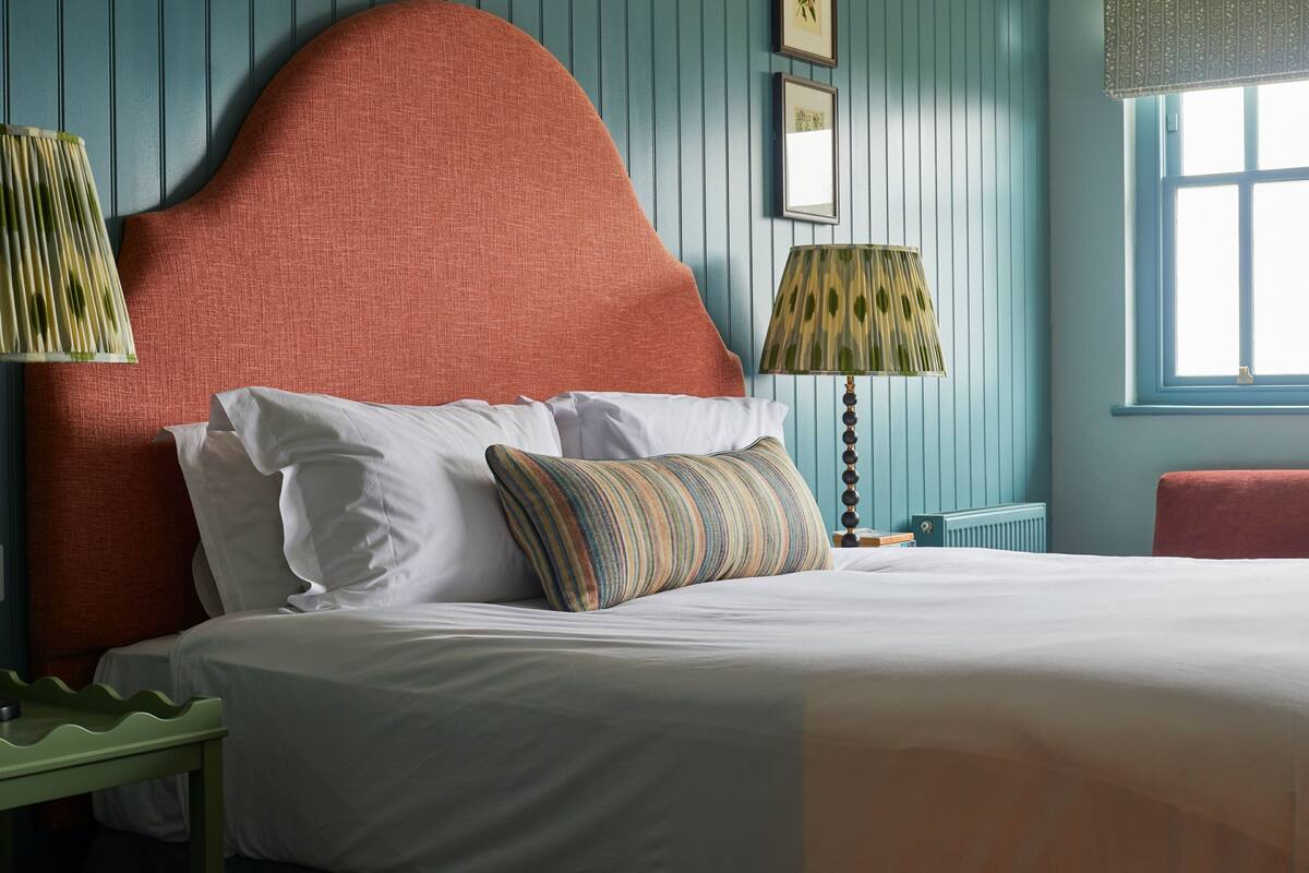 A neatly made bed with white linens and a striped pillow, a green nightstand, and a red upholstered headboard in a room with blue walls and a window.