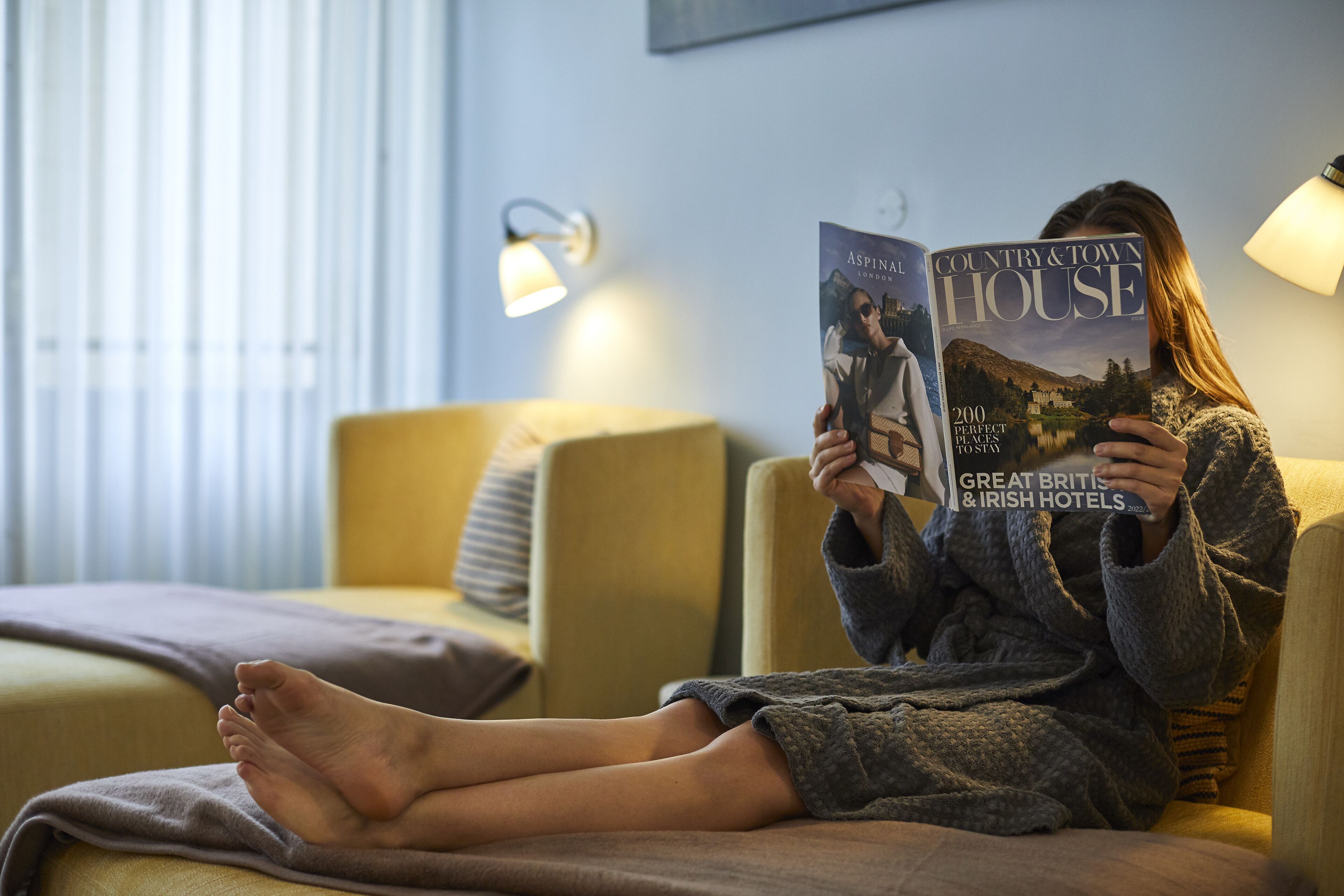 A person sitting on a sofa reading a magazine in a room with a bed, a lamp, and a window with blinds.