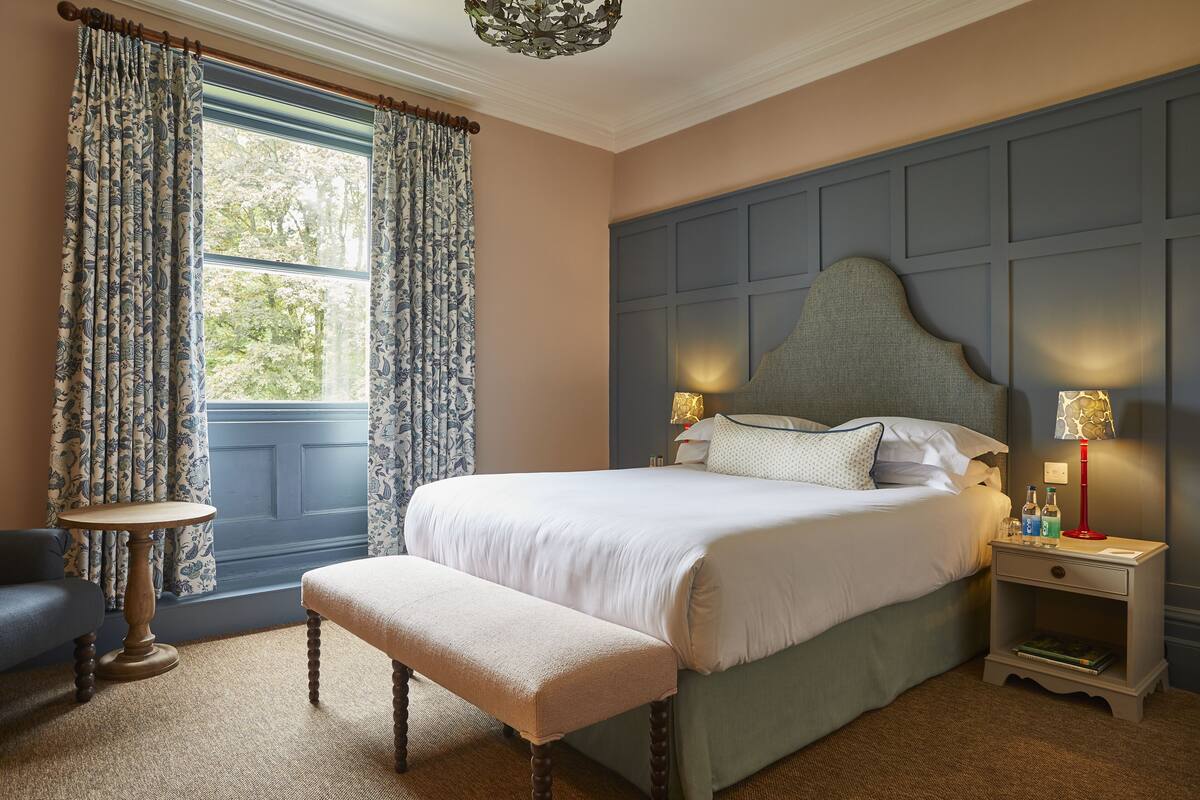 A bedroom with a large bed, a bedside table with lamps, a bench, and a window with curtains.