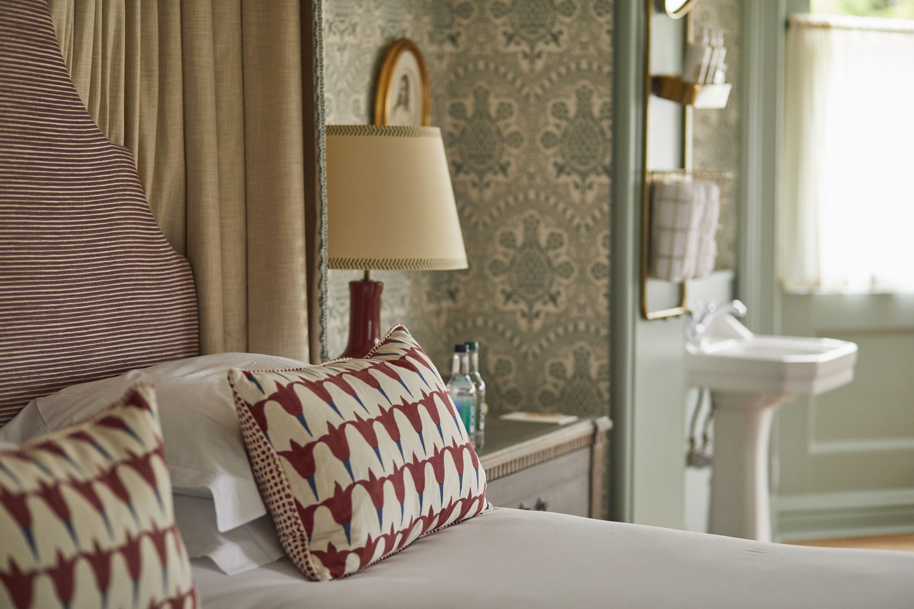A hotel room with a bed, pillows, a nightstand with a lamp, a mirror, a sink, and a patterned wallpaper.