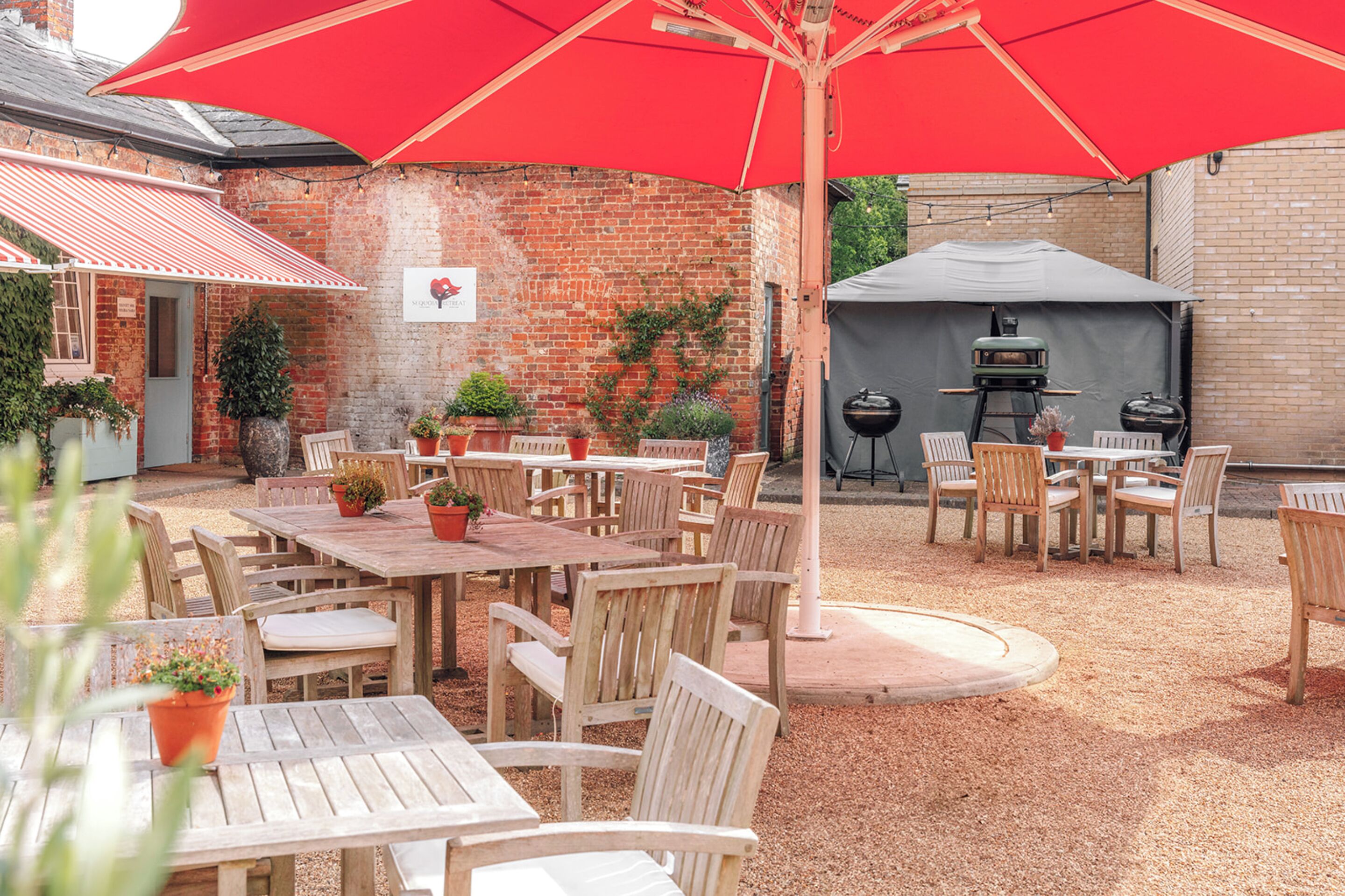Un patio al aire libre con mesas y sillas de madera, un paraguas rojo y una parrilla para barbacoa.