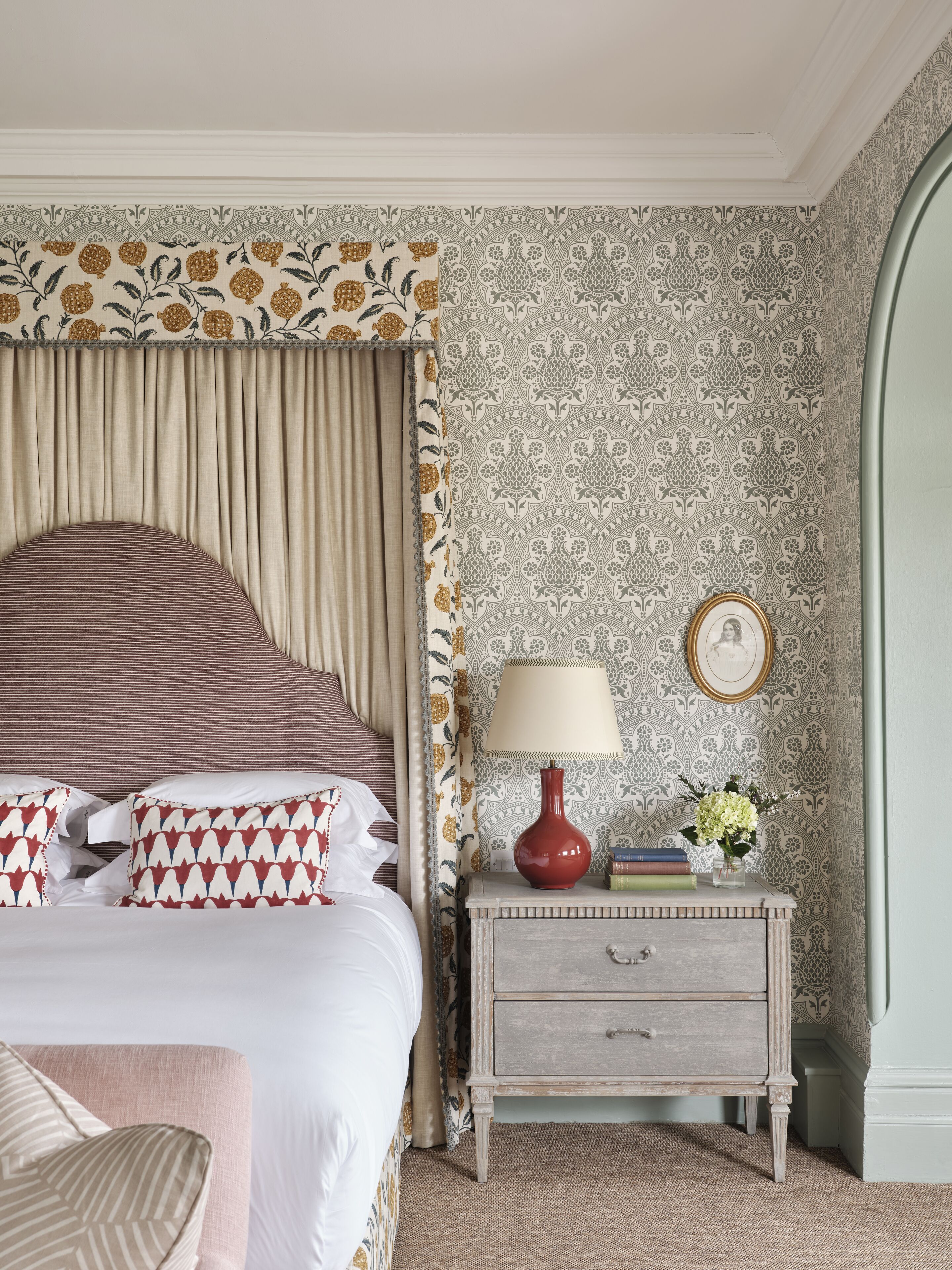 A bedroom with a bed, curtains, a nightstand with a lamp, books, and a vase of flowers.