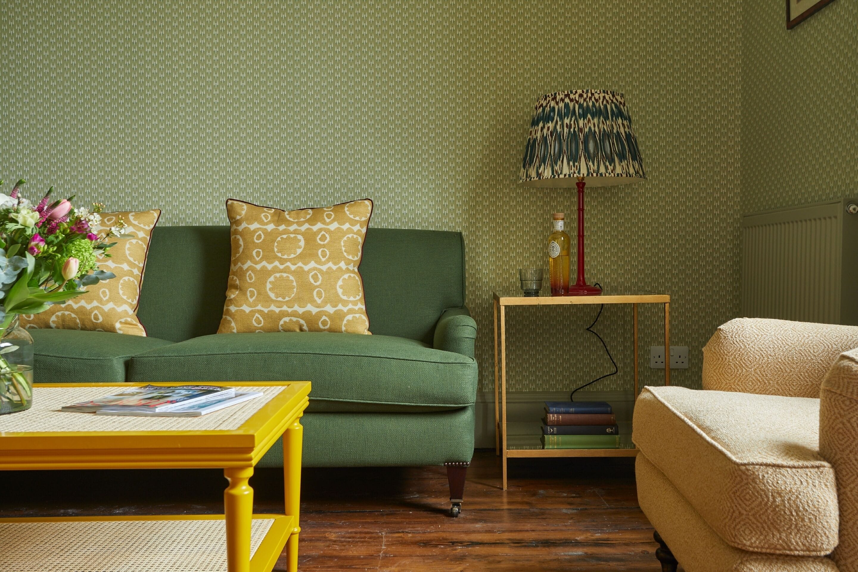 A green sofa with patterned cushions, a small wooden side table with books, a lamp, and a yellow coffee table with magazines.