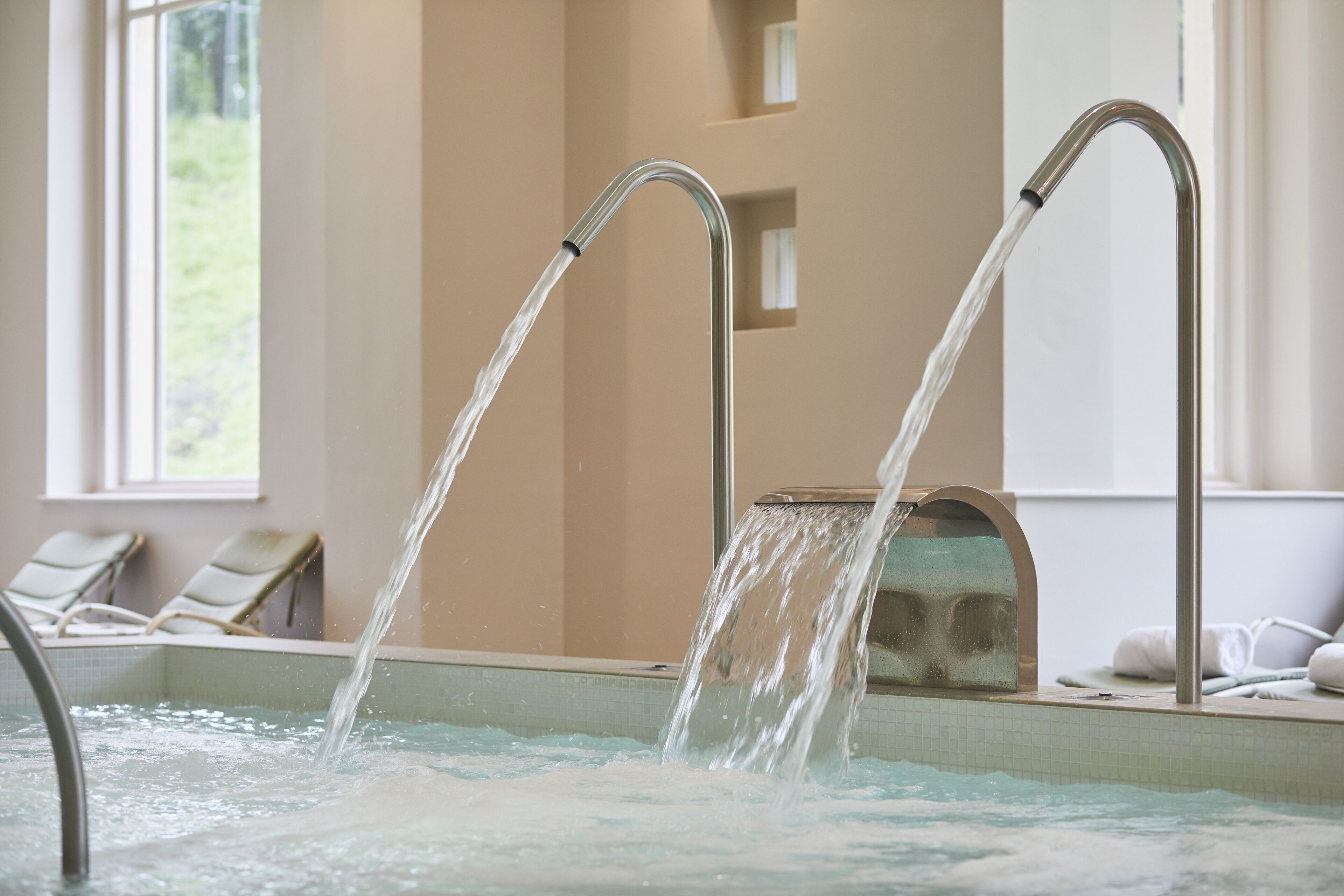 A spa pool with two water jets, lounge chairs, and a view of greenery outside.