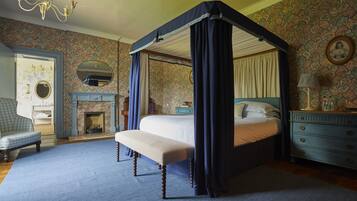 Un lit à baldaquin à quatre poteaux, agrémenté d’un banc, d’une cheminée, d’un fauteuil bleu et d’une commode avec un miroir.
