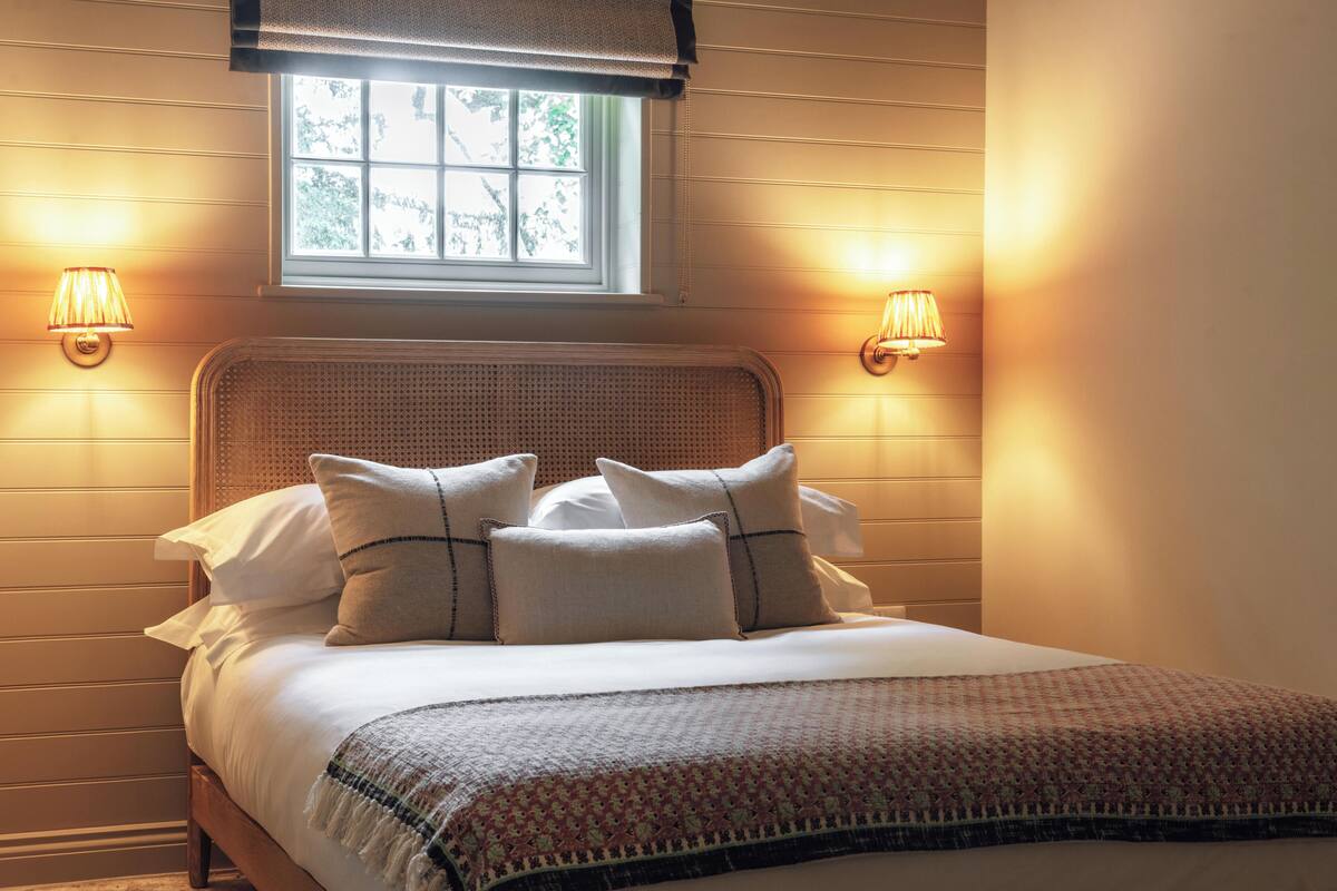 A neatly made bed with a patterned throw, two wall-mounted lamps, and a window with a view of greenery.