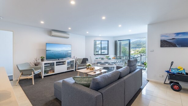A modern living room with a grey sofa, a flat-screen TV, a dining area with a table and chairs, and a balcony with a view of the sea. - Dolphin Cove 8, 2-6 Government Rd - Air conditioning, Wi-Fi, lift, secure parking and close to town (Nelson Bay)