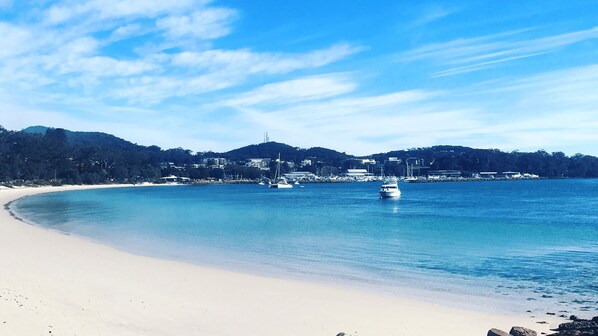 Une plage avec une eau d’un bleu limpide, un rivage sablonneux et quelques bateaux à l’horizon.