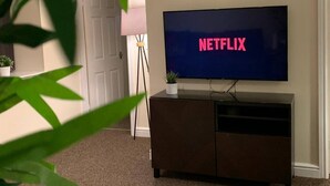 A living room with a television displaying 'NETFLIX' in pink letters, a dark brown entertainment unit, a white door, and a floor lamp. - "the Old Forge"- 5 Minutes Away From Motorway (Bradford)