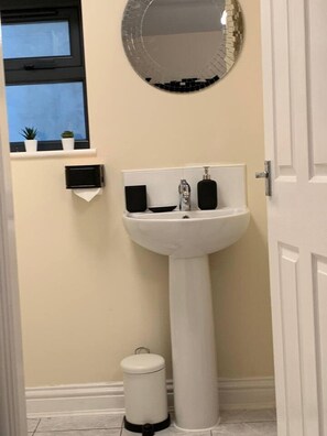 Un baño con un lavamanos blanco de pedestal, un espejo encima y un pequeño cubo de basura al lado.