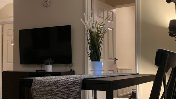 A living room with a flat-screen TV on a stand, a potted plant, and a dining area with a chair and table. - "the Old Forge"- 5 Minutes Away From Motorway (Bradford)