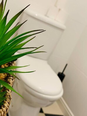 Eine Toilette mit weißem Sitz und Tank, einem schwarzen Spülhebel und einem aus Bändern gewobenen Korb mit grünen Blättern auf der linken Seite.