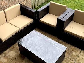 A sectional sofa with beige cushions and a dark wicker frame, placed on a tiled patio with a brick wall and a wooden fence in the background.