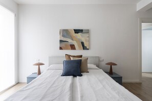 Ein Schlafzimmer mit einem Bett, zwei Nachttischen, einem Wandbild im abstrakten Stil und Blick auf einen Flur.