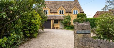A stone house with a gravel driveway leading to a wooden door, surrounded by lush greenery.