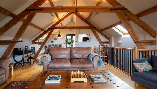 A loft-style living room with exposed wooden beams, a brown leather sofa, a wooden coffee table with chess pieces, and a blue armchair. - Granary at Rectory Farm - sleeps 5 guests  in 3 bedrooms (Shipston-on-Stour)