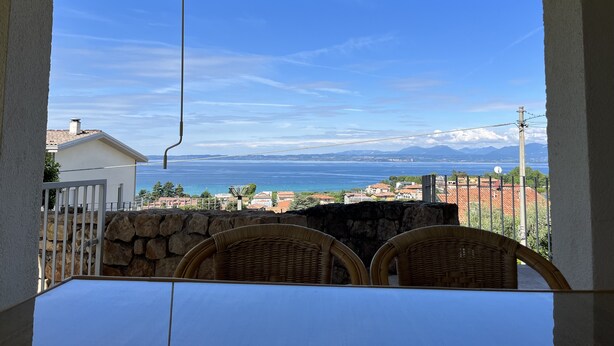 A terrace with a table and chairs, overlooking a coastal view with houses and mountains in the distance. - Apartment with garden and wonderful lake view (Bardolino)