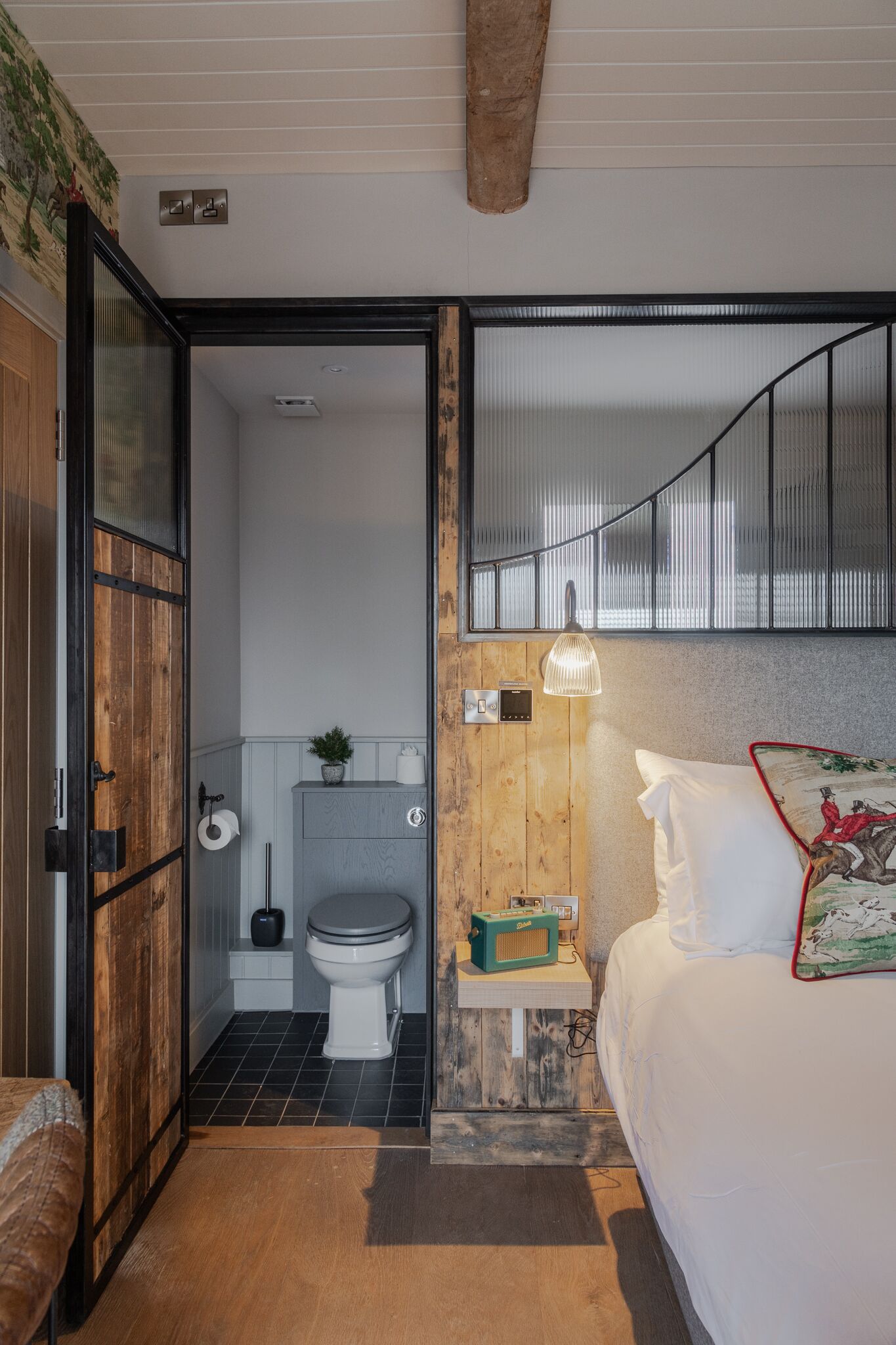 Una camera da letto con un letto, un comodino con una radio, una finestra con persiane e una porta in legno che dà su un bagno con il water e il lavandino.