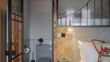 Una camera da letto con un letto, un comodino con una radio, una finestra con persiane e una porta in legno che dà su un bagno con il water e il lavandino.