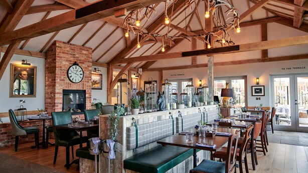 A restaurant with wooden beams, a brick wall, and a clock. Tables and benches are arranged for dining. - Hare & Hounds Newbury (Newbury)