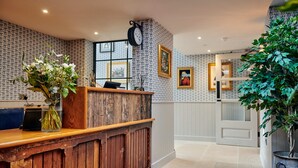 A reception area with a wooden counter, a clock, framed artwork, and a potted plant. - Hare & Hounds Newbury (Newbury)