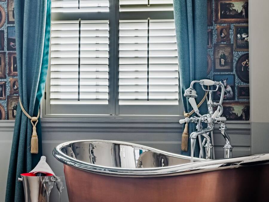 Un baño con una bañera independiente, una ventana con persianas y un papel tapiz estampado.