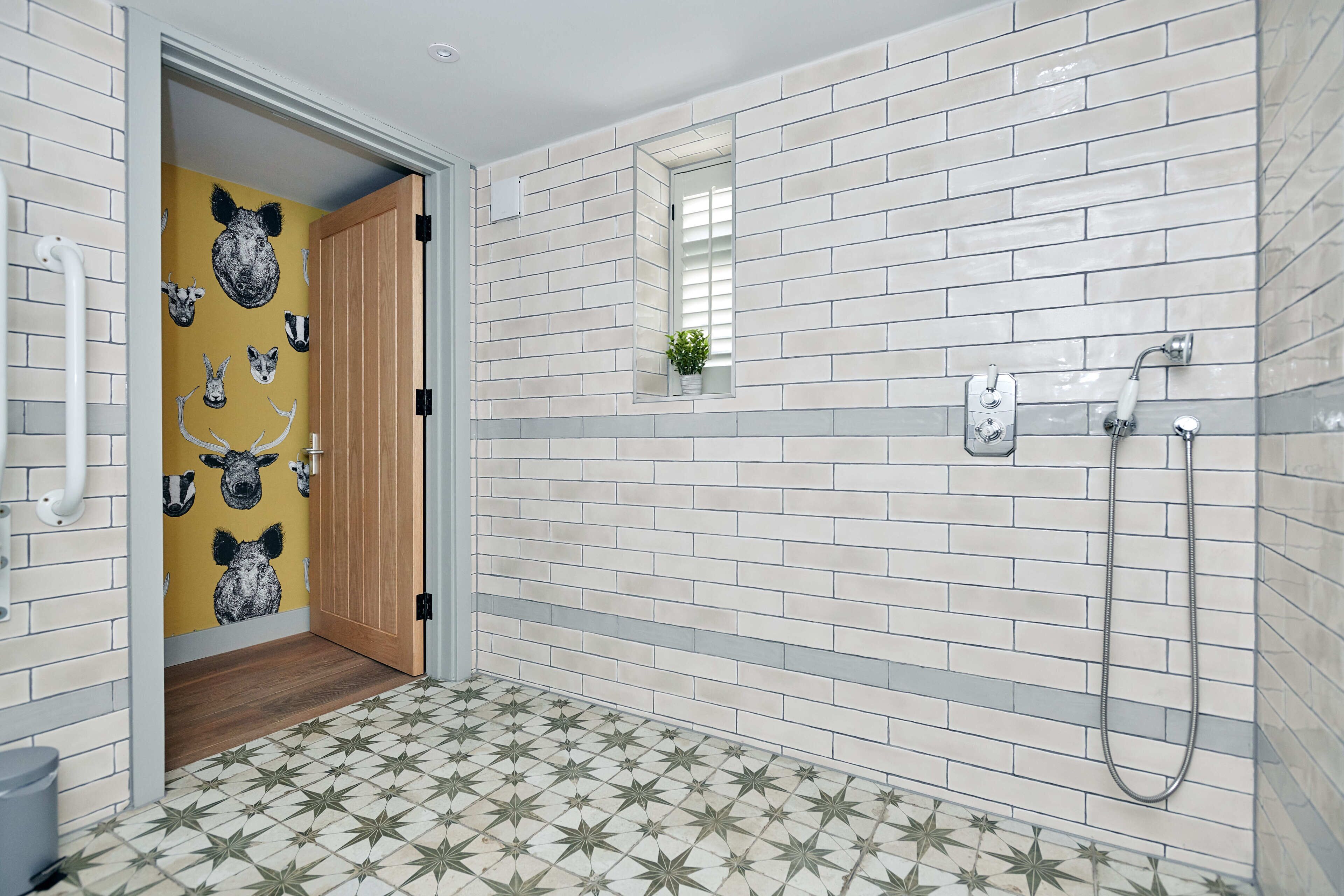 Un baño con azulejos blancos y grises, una puerta de madera y una ducha con cabezal de ducha manual.