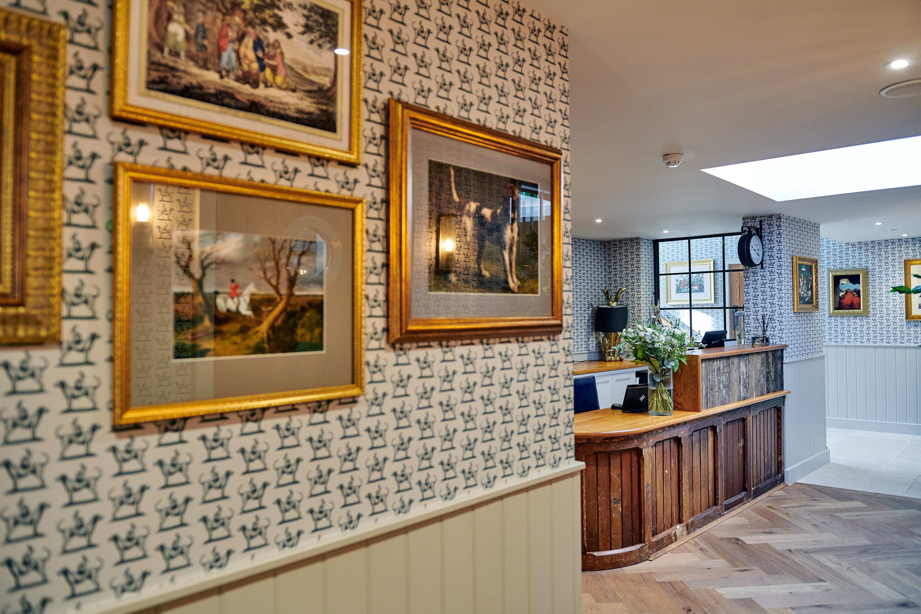 A hotel lobby with patterned wallpaper, framed artwork on the walls, a reception desk, and a wooden floor.