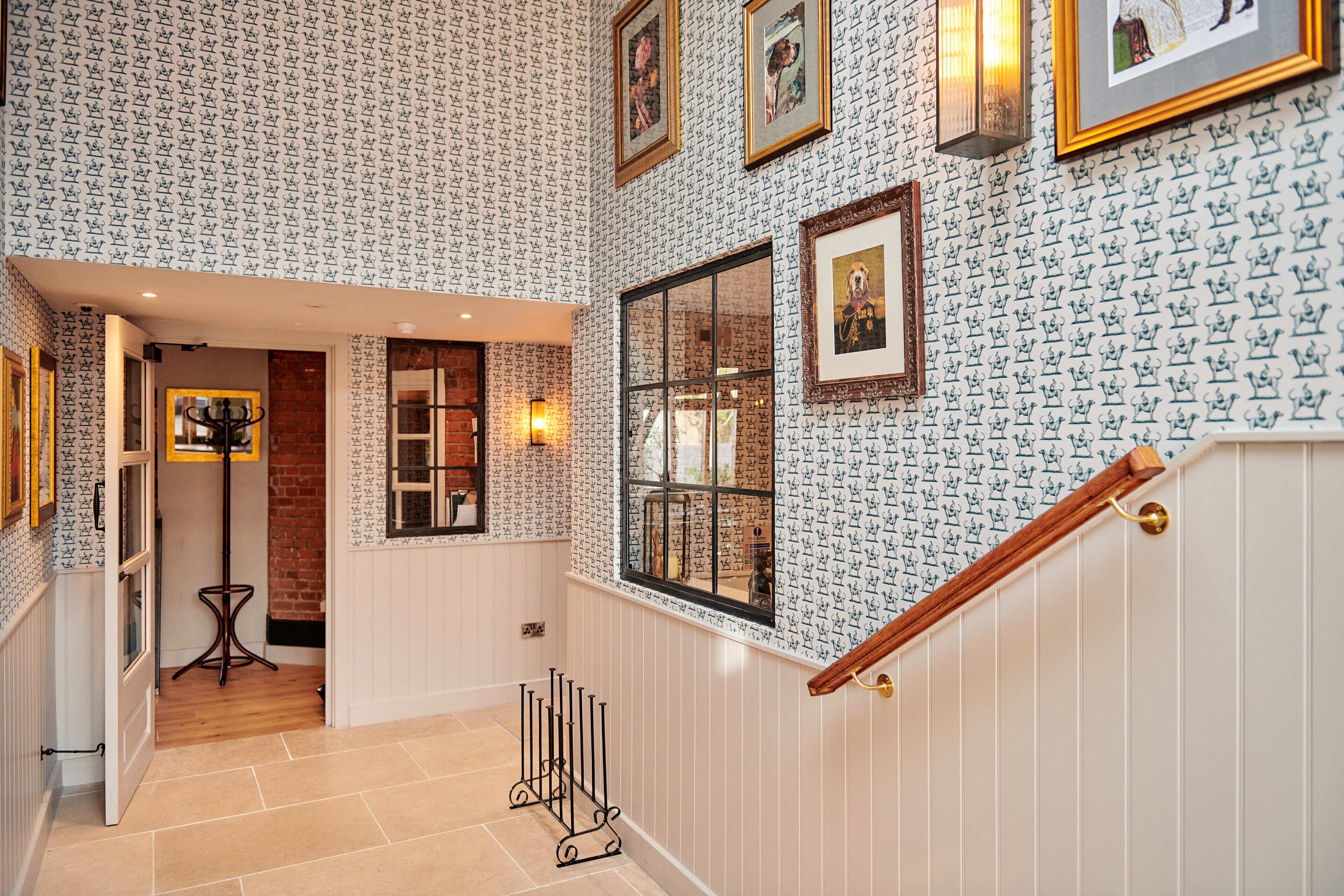 Un pasillo con papel tapiz estampado, obras de arte enmarcadas y una escalera de madera.