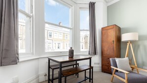 Une pièce avec vue sur les bâtiments, grâce à de grandes fenêtres, un meuble en bois, une chaise avec une lampe et une table sur laquelle est posé un vase.