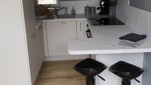 A modern kitchen with white cabinets, a black countertop, and two black bar stools. - Cosy apartment in the heart of Bayeux (Bayeux)