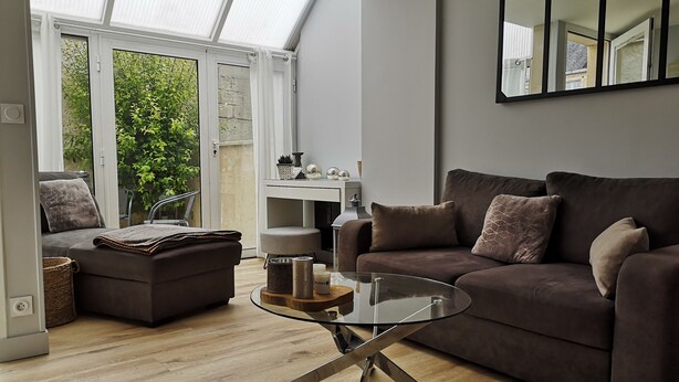 A modern living room with a glass coffee table, a brown sofa, and a large window with curtains. - Cosy apartment in the heart of Bayeux (Bayeux)
