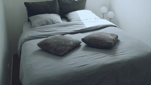A bedroom with a skylight, a bed with grey bedding and two pillows, a bedside table with two white spherical lamps, and a view of a building outside. - Cosy apartment in the heart of Bayeux (Bayeux)