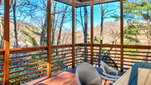 Un terrazzo in legno con vista su alberi e colline. Sono presenti due sedie e un tavolo.