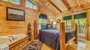 A log cabin bedroom with a large bed, a wooden headboard, a built-in bathtub, and a view of the outdoors. - 3 BR w/ Hot Tub, WiFi, Pool Access, Close to Town, Free Attraction Tickets Daily (Pigeon Forge)