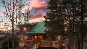 A rustic log cabin with a green roof, a porch, and a staircase leading to the entrance. - 3 BR w/ Hot Tub, WiFi, Pool Access, Close to Town, Free Attraction Tickets Daily (Pigeon Forge)