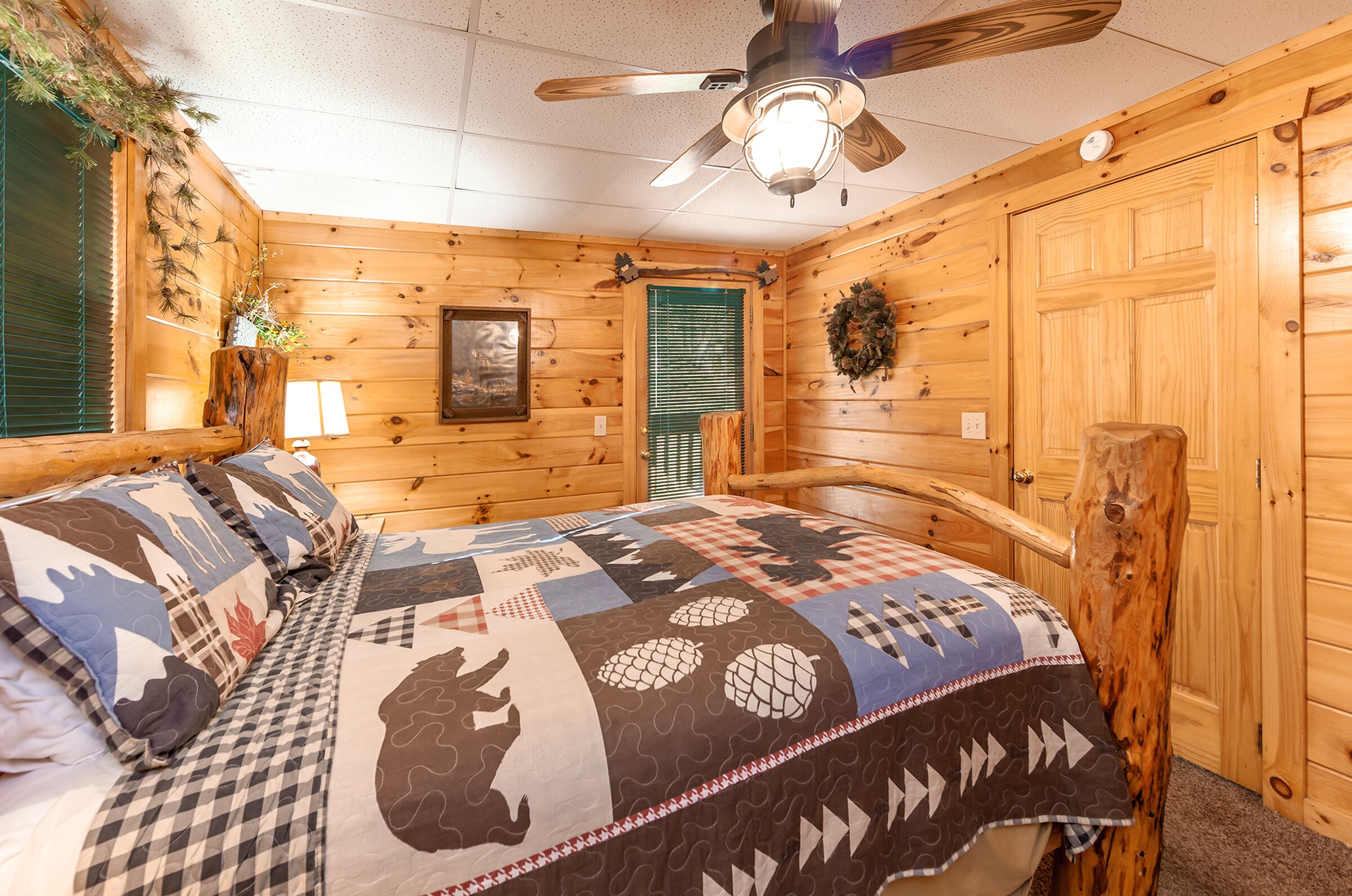 A cozy log cabin bedroom with a large bed, wooden walls, a ceiling fan, and a green window blind.