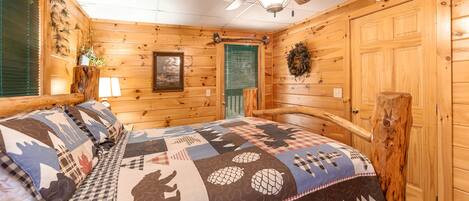 A cozy log cabin bedroom with a large bed, wooden walls, a ceiling fan, and a green window blind.