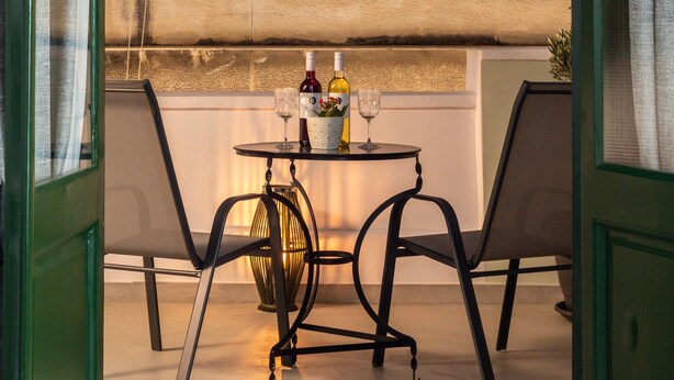 Un balcon ouvert avec deux chaises, une table sur laquelle se trouvent des bouteilles et des verres, et une vue sur un immeuble se dressant sous un ciel sombre.