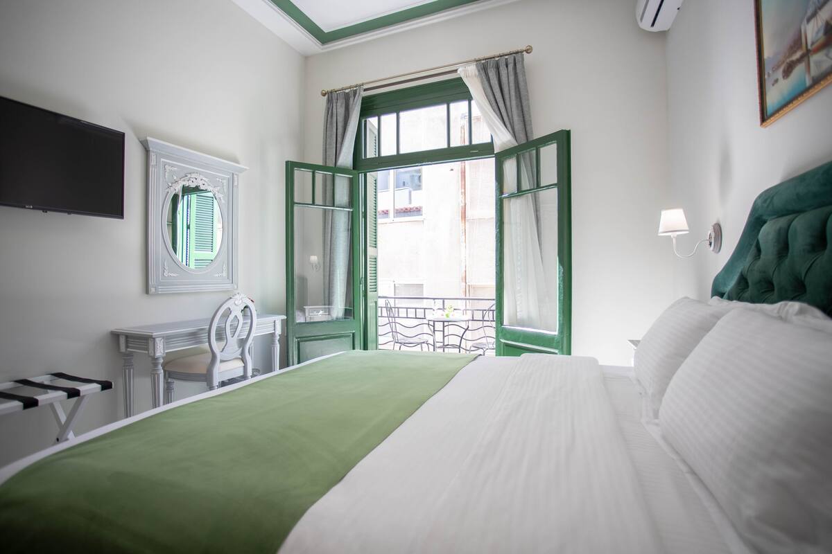 A hotel room with a bed, a desk, a chair, a TV, a window with curtains, and a balcony view.