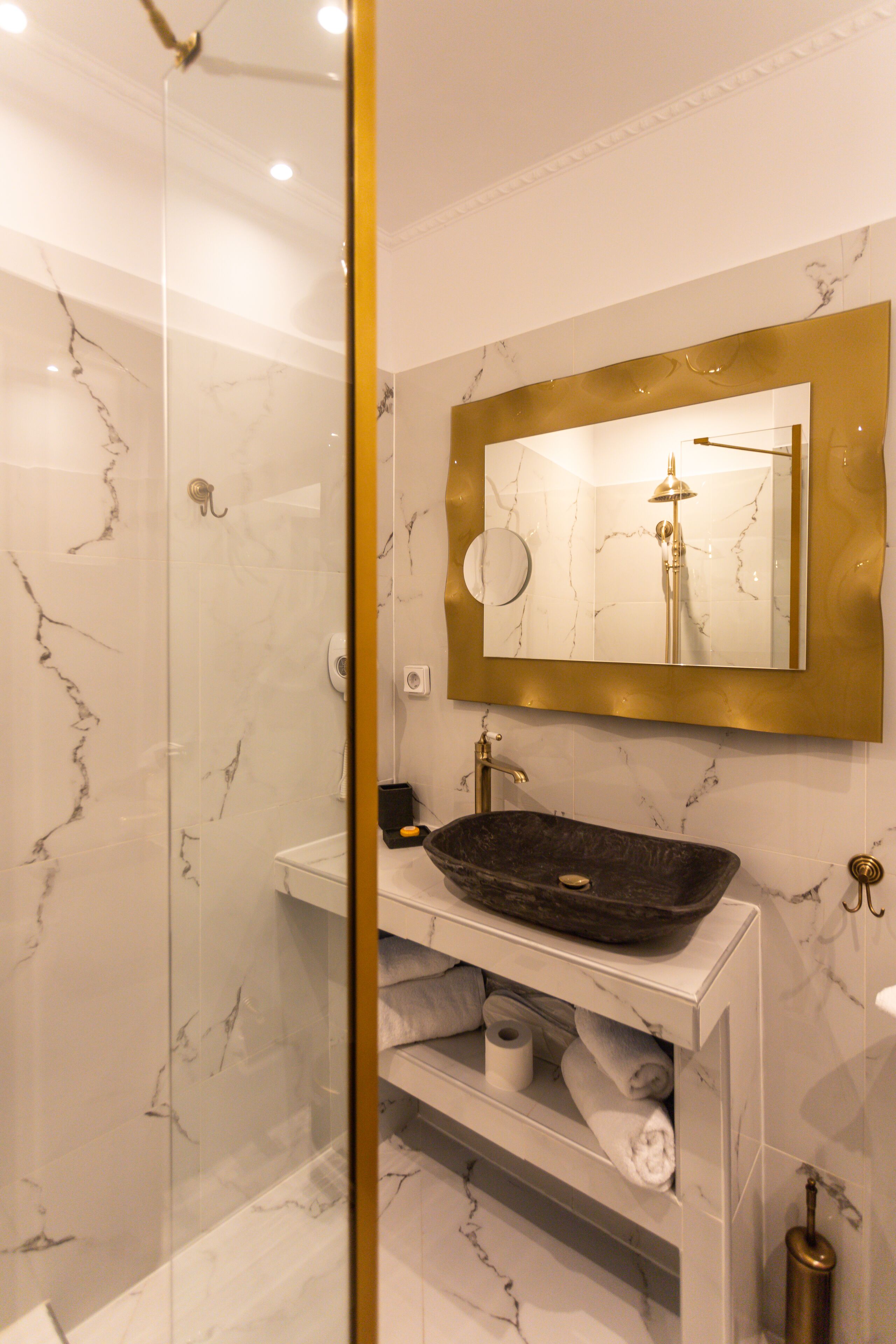 Une salle de bain avec un mur en marbre, un miroir encadré d’or, un lavabo noir et un porte-rouleau de papier toilette.